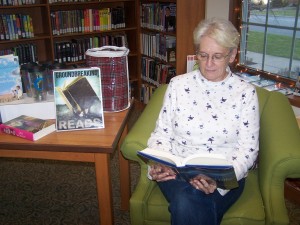 Carol Haab reads one of her favorite books while sitting by the table of prizes available for Milford Public Library's Adult Reading Club winners. Adult Reading Club begins Jan. 2 and lasts until March 30. Sign up at the Adult department desk and you could win one of these prizes!