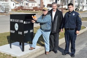 Paul Newcomer, left, president-elect ofWawasee Kiwanis, shows how easy it is to drop off expired or unwanted prescription medication at the new medication drop box on the northside of the Syracuse Town Hall. Wawasee Kiwanis donated funds toward the purchase and installation of the box. Shown to the right are Syracuse Police Chief Tony
Ciriello and officer Michael Bumbaugh. (Photo by Deb Patterson)