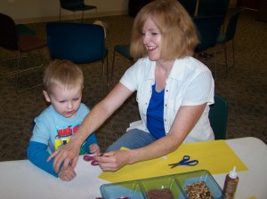 Patty Kratzer helps her grandson, Lukas Kratzer, cut  out a sunflower at story time. The theme was seeds and the children made sunflowers by gluing seeds in the middle of a yellow flower. These flowers are on display by the South door of the Milford Public Library.   (Photo provided) 