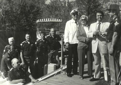 Shown during what is believed to be the donation of a lake patrol boat. Shown in the boat are lake patrol officers Warren Swartz, Don McCulloch, Joe Thornburg, Terry McCarty and Tom Kitch. On shore the only individuals known are Jane Nearing and Sheriff Al Rovenstine, far right.
