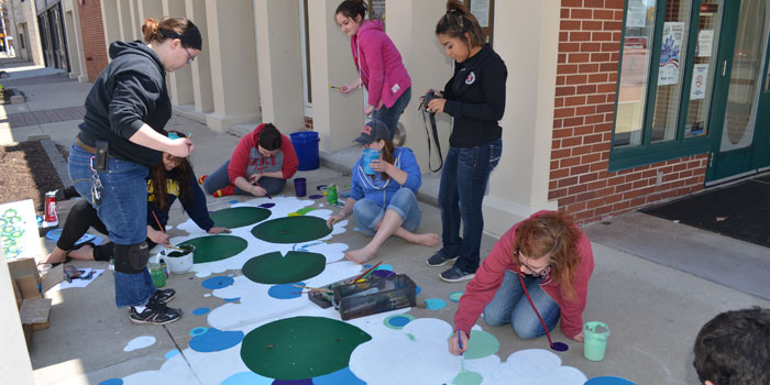 Students from Grace College paint a mural titled, "Lily Pad Hopscotch" on the sidewalk outside the Kosciusko Chamber of Commerce building. (Photo by Amanda McFarland)