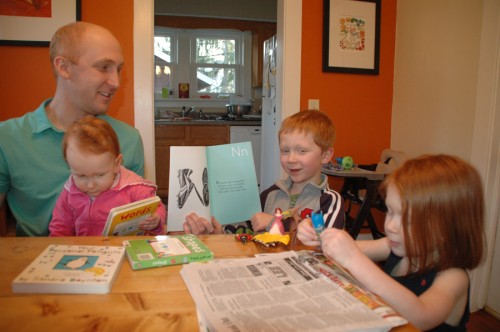 SARAH’S LITTLE HELPER — The Steele's son, 5-year old Corban, second from right, reads a passage from “The Shoephabet.” It’s about his favorite sport, baseball. Corban often runs the Power Point  when his mom gives presentations of the book at schools and libraries. Sister Jaeda is at right and Liddi, 1, sits on James’ lap. (Photo by David Hazledine)