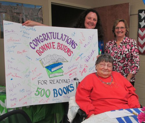 Pam Long and Debbie Conner of the North Webster Library (standing) present a giant card signed by well-wishers to Bonnie Burns in recognition of her achievement of reading 500 books. Working to overcome reading challenges, Burns is one of the library’s most avid readers.